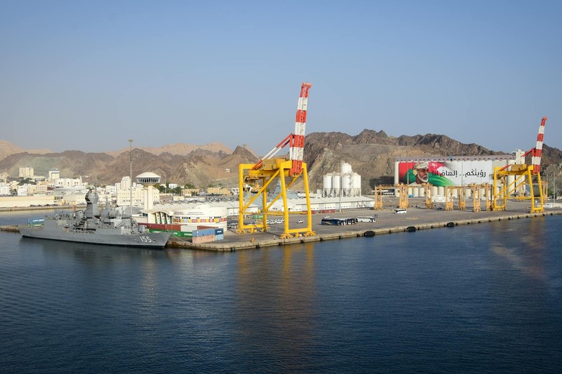 Aerial view of a Middle Eastern port with cargo cranes, storage tanks, and a moored naval vessel against a backdrop of arid mountains