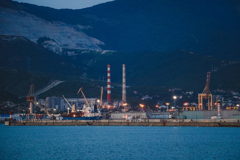 Industrial facilities with smokestacks and cranes along a body of water at dusk