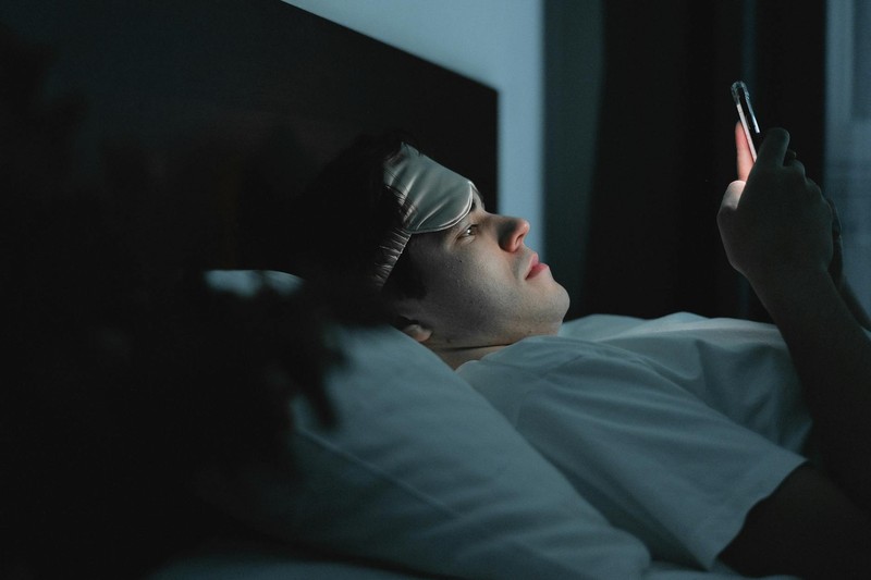 A person lying in bed at night illuminated by a smartphone screen, wearing a sleep mask pushed up