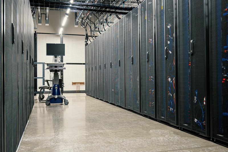 Rows of black server racks in a modern data center with overhead cabling and fluorescent lighting
