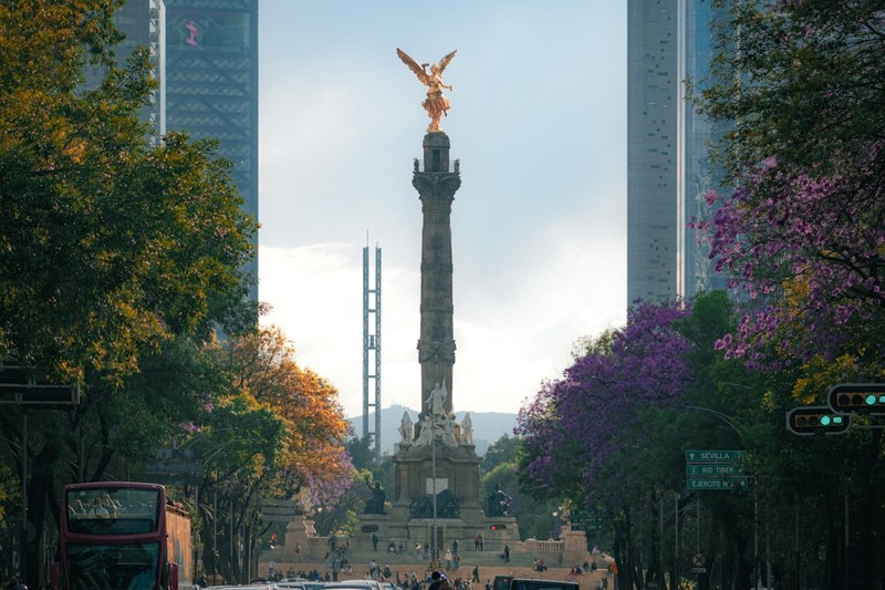 The Angel of Independence monument in Mexico City, with a tall gilded angel statue atop a column surrounded by trees and modern skyscrapers
