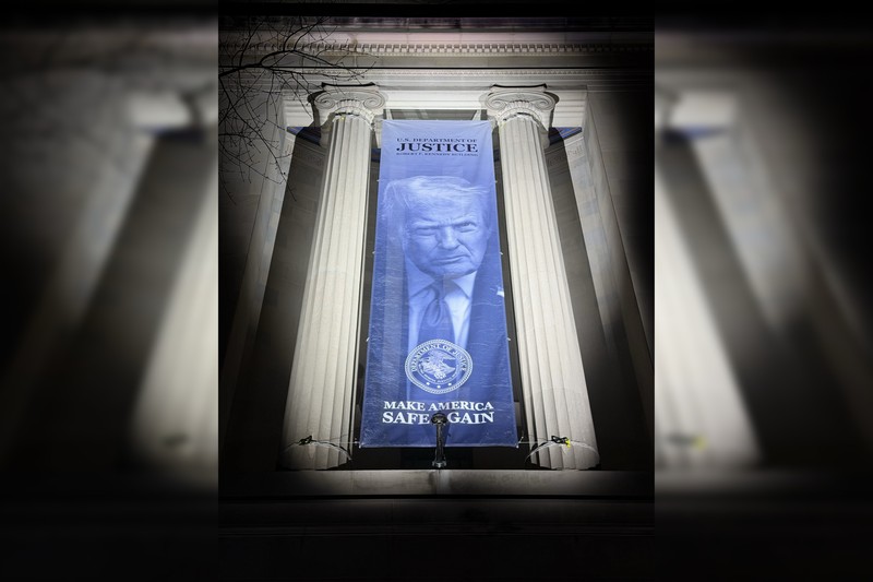 A banner displaying Donald Trump's portrait and the words 'U.S. Department of Justice' hangs between the columns of the DOJ headquarters building in Washington, D.C.