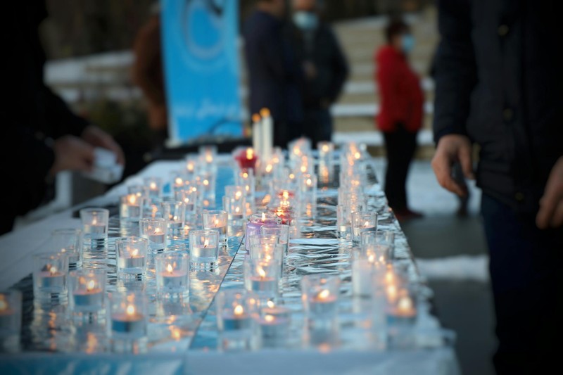 Lit candles arranged on a white tablecloth at an outdoor candlelight vigil with mourners in the background