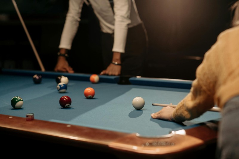 Two players in a dimly lit pool hall focusing on a billiards shot on a green felt table