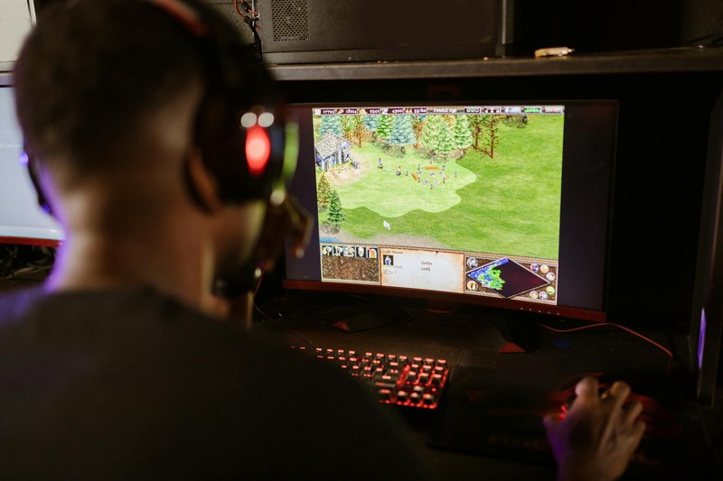 A gamer seen from behind plays a retro-styled video game on a monitor in a dimly lit room