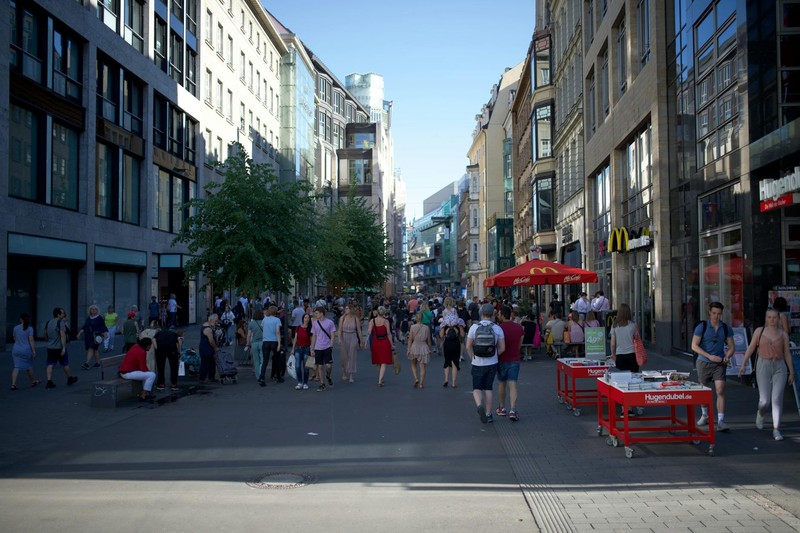 A bustling pedestrian shopping street in Leipzig, Germany lined with multi-story buildings, outdoor cafés, and pedestrians on a sunny day.