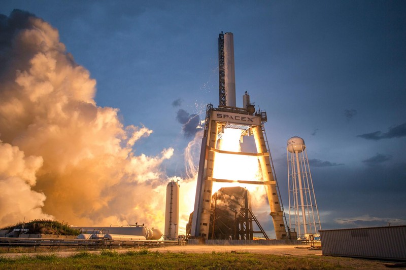A SpaceX rocket launches from a pad at dusk, with bright flames and exhaust illuminating the launch tower against a twilight sky.