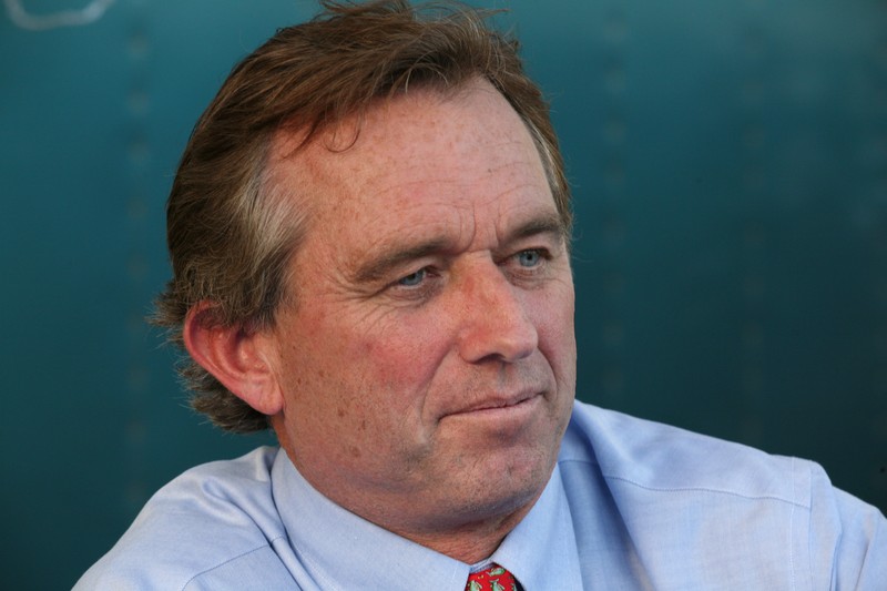 Robert F. Kennedy Jr. in a professional portrait wearing a light blue shirt and red tie against a teal background