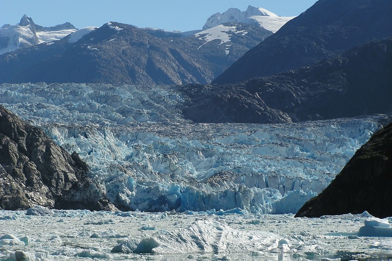 A tidewater glacier fills an Alaskan fjord, framed by steep snow-capped mountains with broken ice floating in the foreground