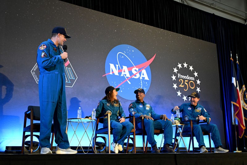The four Artemis II crew members in blue NASA jumpsuits on stage at a NASA event with the Freedom 250 emblem behind them