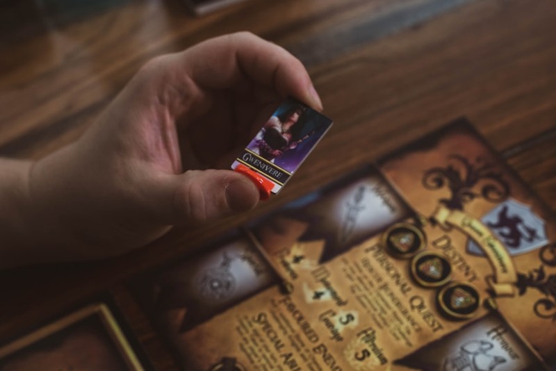 A hand holds a fantasy game card above an ornate medieval-themed game board on a wooden table