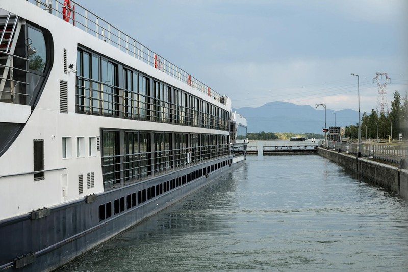 A multi-deck cruise ship navigating a narrow waterway with a scenic landscape in the background