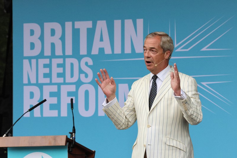 Nigel Farage gestures while speaking at a Reform UK political rally, with a large blue backdrop reading 'BRITAIN NEEDS REFORM'