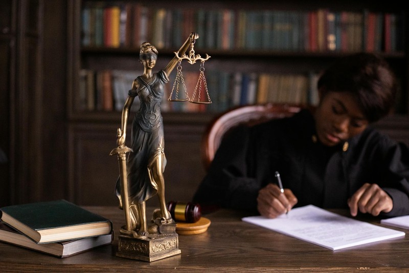 A judge in black robes writes at a wooden desk beside a Lady Justice statue and gavel in a law office