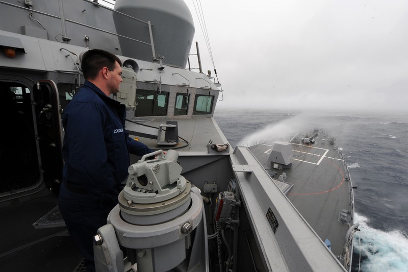 A U.S. Navy warship transits through choppy seas under an overcast sky, with a sailor standing watch on deck.