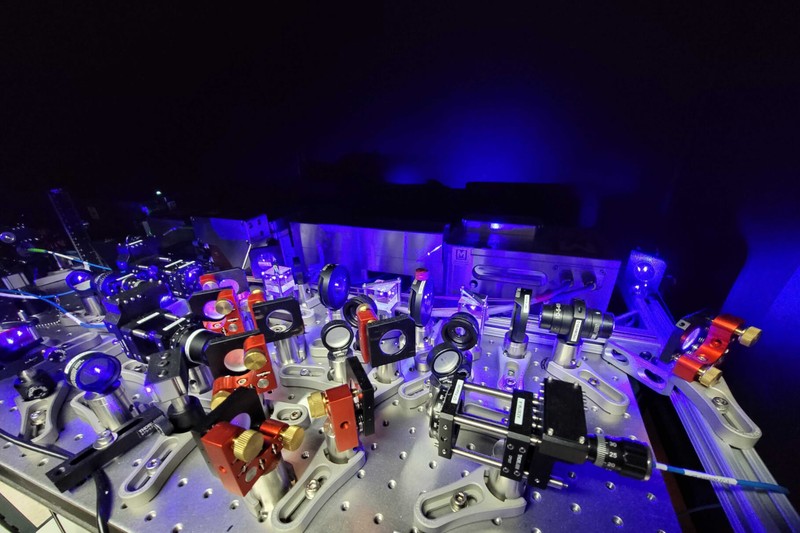 A dense optical table covered with lasers, lenses, mirrors, and precision mounting equipment in a blue-lit quantum computing laboratory