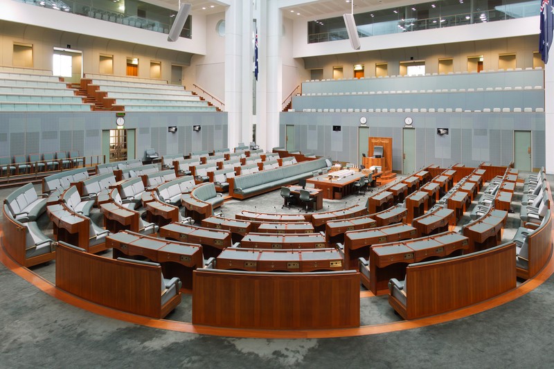 Empty Australian House of Representatives chamber in Parliament House, Canberra, with semicircular seating and polished wooden desks.