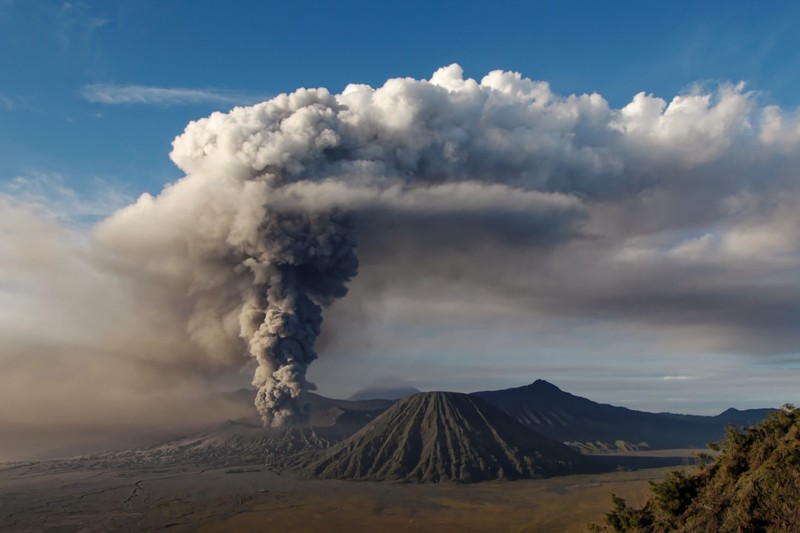 A volcano erupting with a large ash plume rising into the sky above a barren volcanic landscape