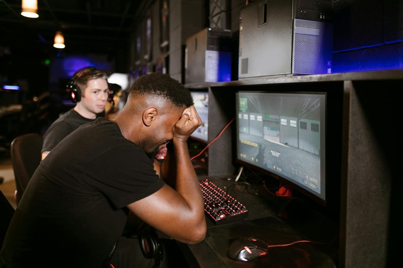 Two men at gaming PCs in a dark gaming setup, one appearing frustrated with hand on forehead