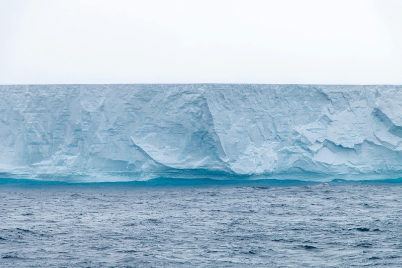A massive blue tabular iceberg floats in dark Antarctic waters under an overcast gray sky