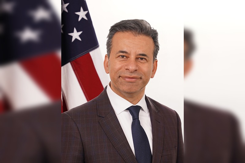 Official portrait of FDA Commissioner Dr. Marty Makary wearing a plaid suit and blue tie, with an American flag visible in the background