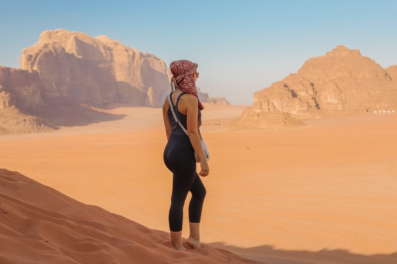 A person stands atop a sand dune gazing across a vast desert landscape with towering rock formations under a clear sky