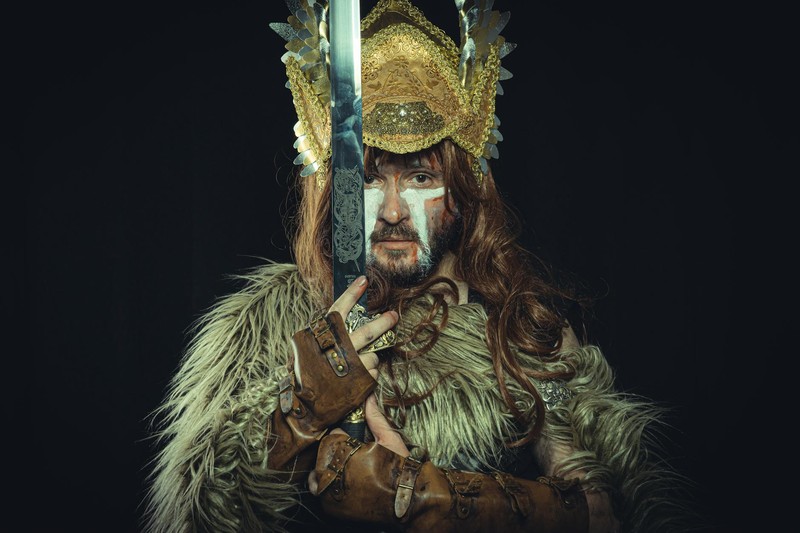 A warrior in ornate armor and fur garments holding an engraved sword against a black background