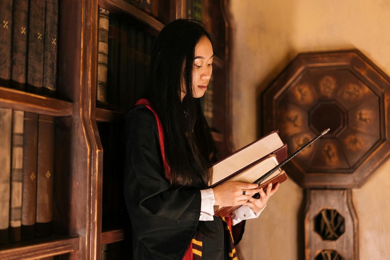 A young woman in a wizard robe studies a book in a warm, vintage library filled with leather-bound volumes