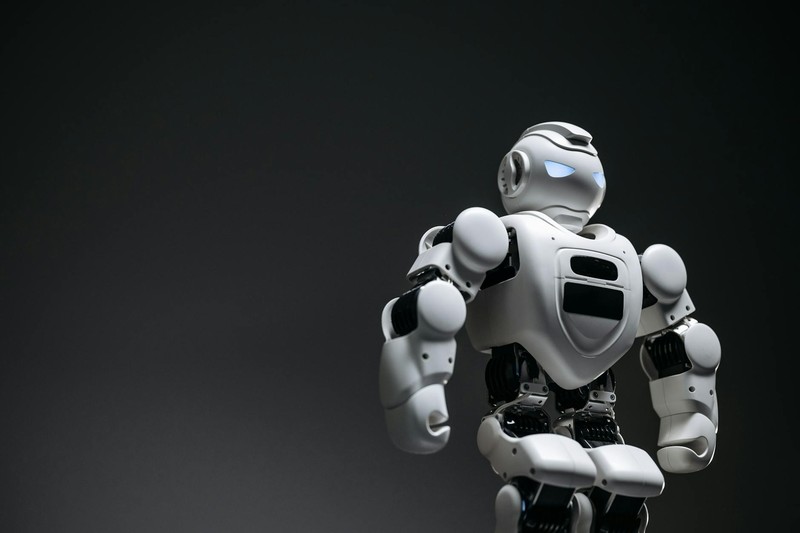 A white humanoid robot with a blue illuminated sensor eye stands against a dark gradient background in a studio shot