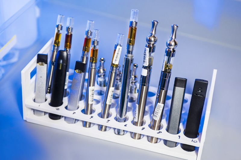 Assorted e-cigarette and vaping devices arranged in numbered test tube slots for laboratory analysis