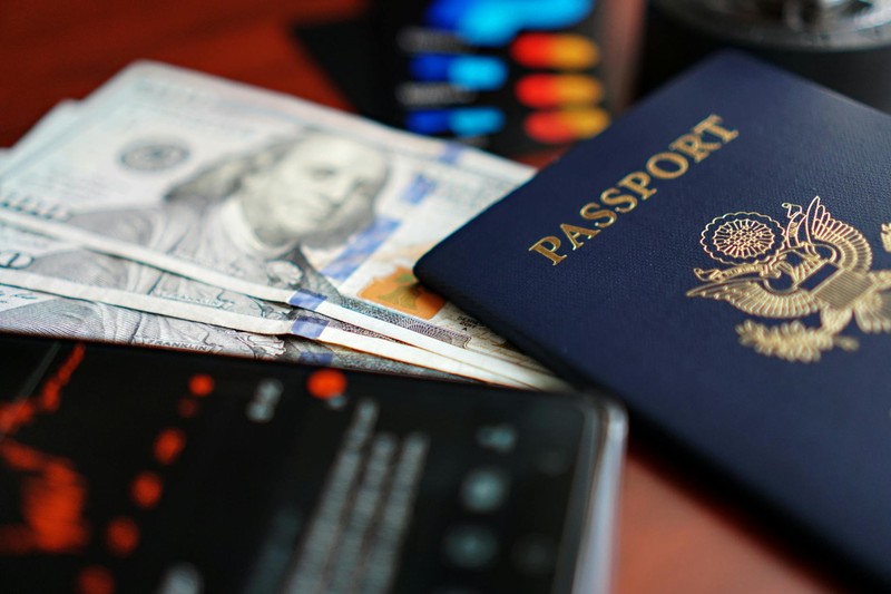 A US passport and fanned hundred-dollar bills arranged on a table beside a smartphone displaying financial data