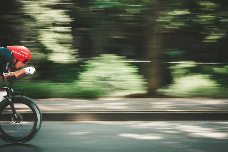A cyclist races at high speed along a road, captured with dramatic motion blur emphasizing velocity