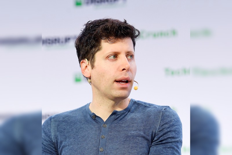 Sam Altman speaking onstage at a conference, wearing a blue shirt with an earpiece microphone