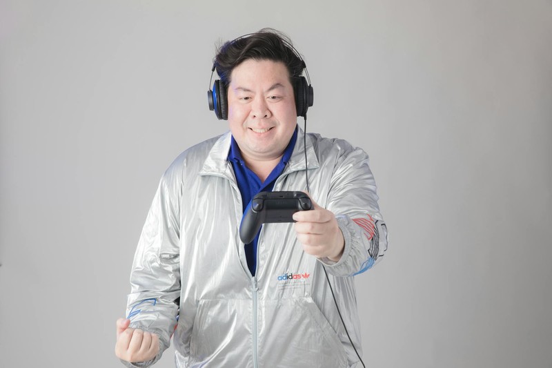 Asian man wearing headphones and silver jacket holding a game controller with an excited expression