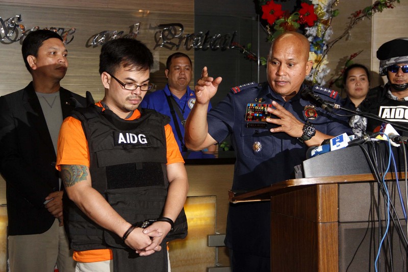 Philippine National Police Chief Ronald Dela Rosa speaking at a press conference podium alongside a handcuffed suspect