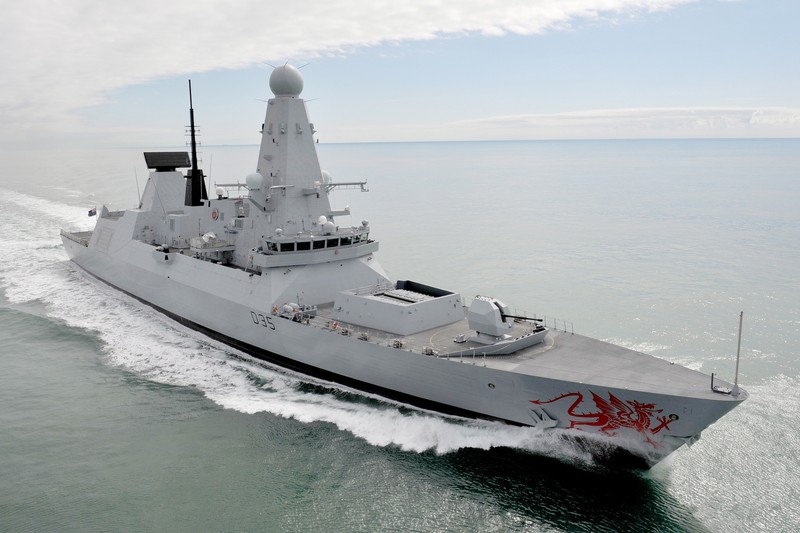 A Royal Navy Type 45 destroyer cuts through open ocean with a visible wake, its radar dome and angular superstructure silhouetted against a cloudy sky.