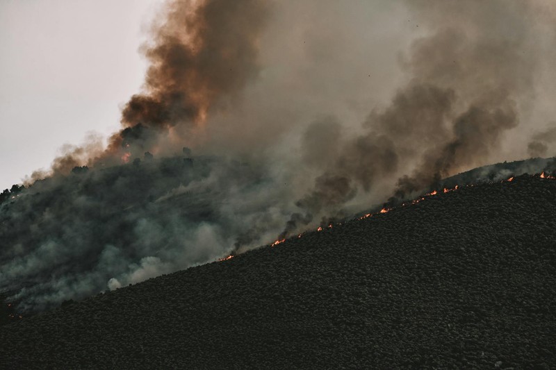 Intense wildfire burning across a darkened mountainside with thick smoke billowing into the sky