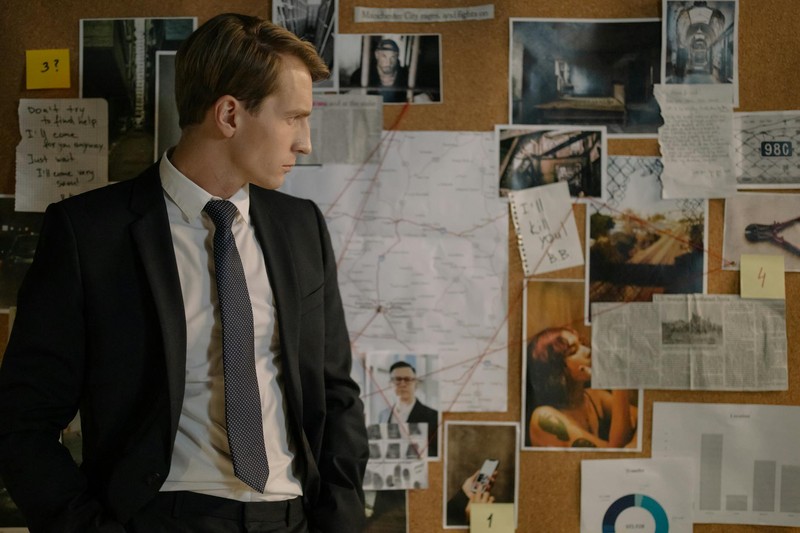 A detective in a dark suit examines a corkboard covered with evidence photos, maps, and notes