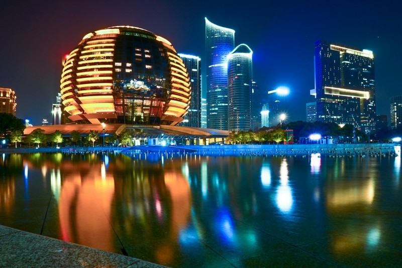 Nighttime panorama of Hangzhou's illuminated modern skyline reflected in calm water, featuring a distinctive spherical building surrounded by sleek skyscrapers