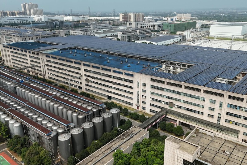 Aerial view of a modern industrial manufacturing facility with solar panels on the roof, cylindrical storage tanks, and surrounding infrastructure near an urban area.