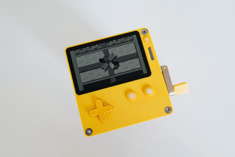 A bright yellow Playdate handheld gaming console with a grayscale screen, D-pad, and two white buttons, displayed on a white background