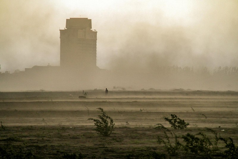 A thick dust storm engulfs a landscape with a building and sparse vegetation, reducing visibility to near zero
