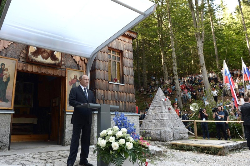 Vladimir Putin speaks at a memorial ceremony near a wooden chapel, with Russian flags visible in the crowd.