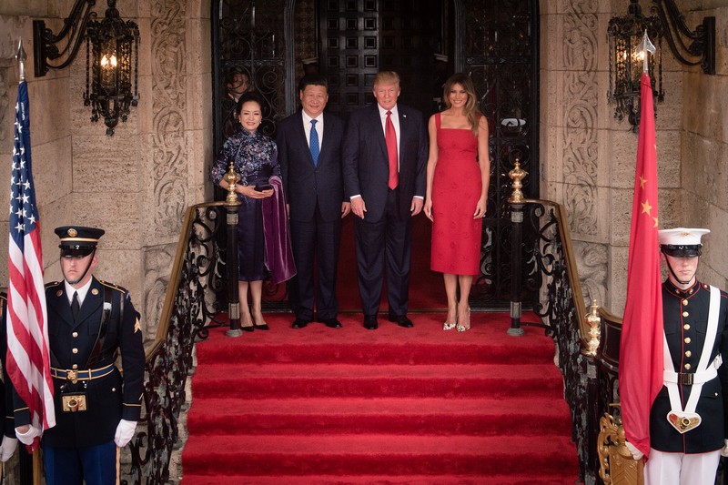 Donald Trump and Xi Jinping with their spouses standing on a red-carpeted staircase flanked by U.S. and Chinese flags during a formal diplomatic welcome