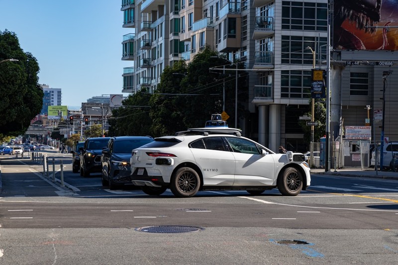 A white Waymo Jaguar I-Pace robotaxi with roof-mounted sensors at a city intersection