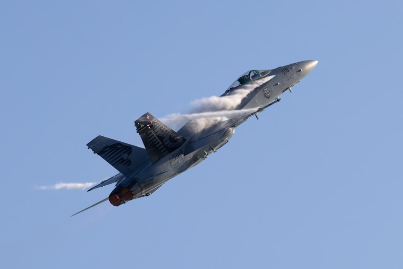 An F/A-18 Hornet fighter jet climbs steeply through a clear blue sky with vapor trails and afterburner glow
