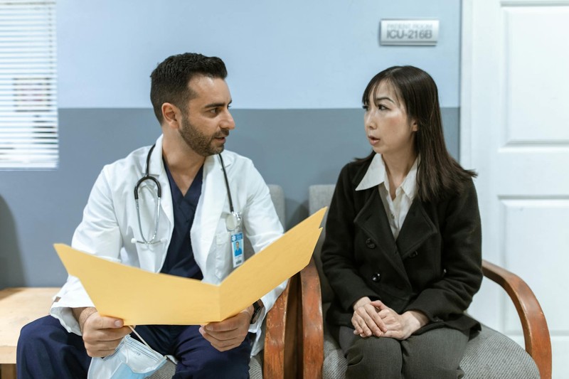 A doctor in a white coat reviews a patient chart during a medical consultation with a woman in a clinical setting.