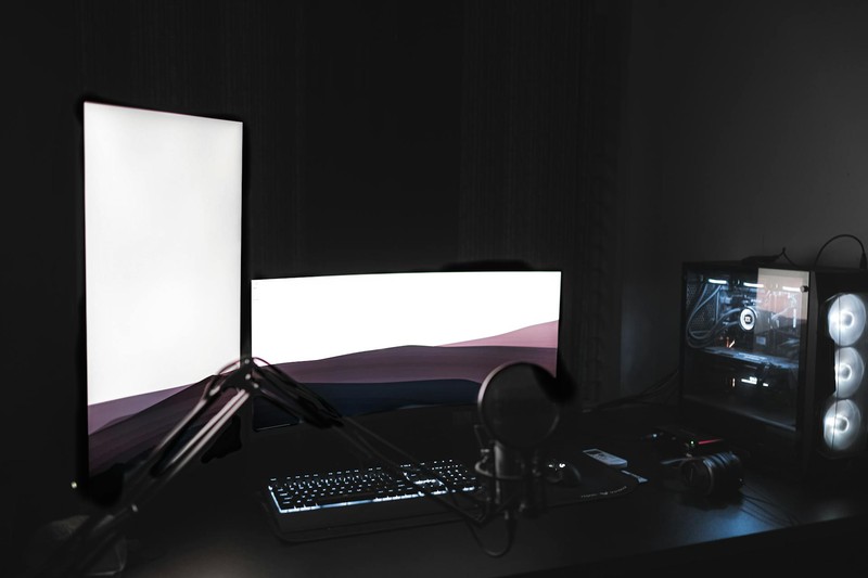 A dark gaming setup with dual monitors, RGB-lit PC tower, mechanical keyboard, and streaming microphone