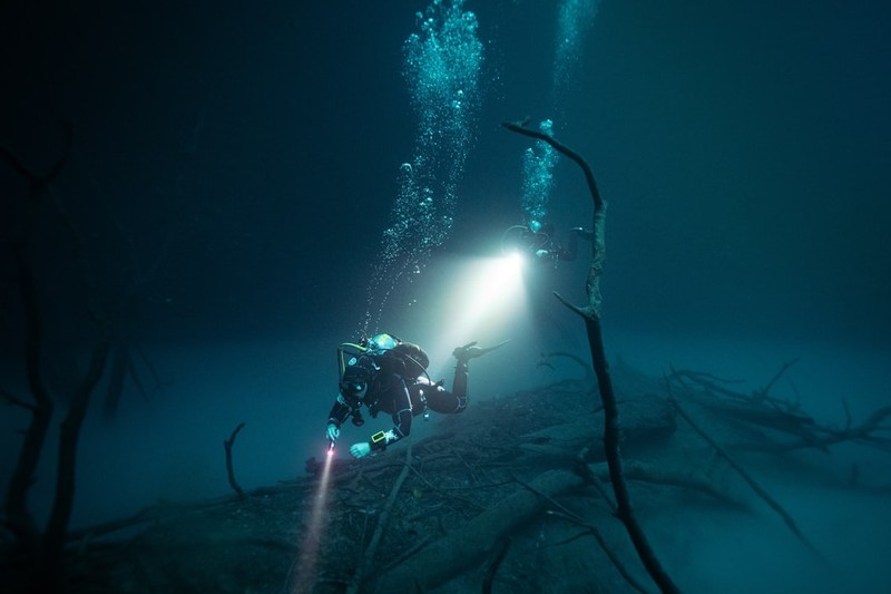 A lone scuba diver with a flashlight explores a dark, mysterious underwater cenote