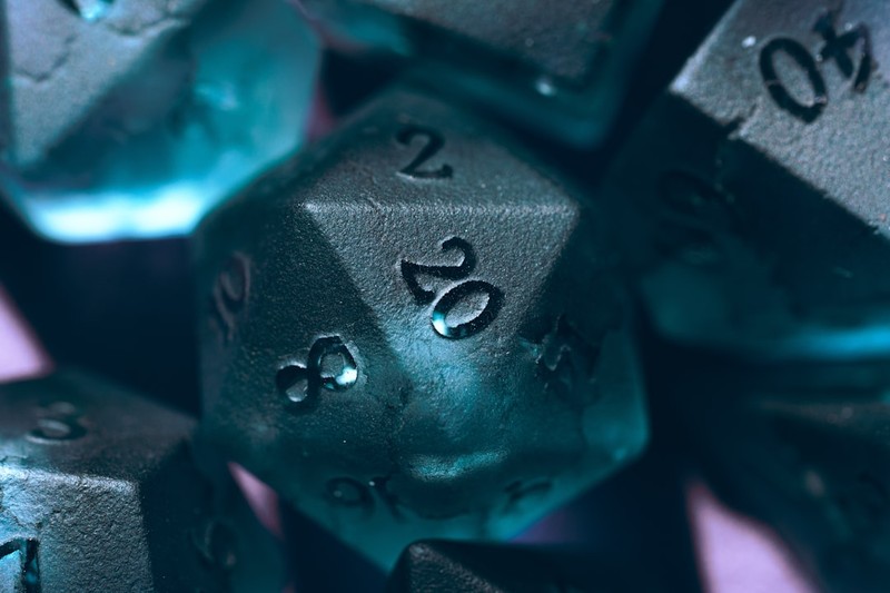 A teal d20 polyhedral gaming die with the number 20 facing up, illuminated by dramatic blue lighting against a dark background